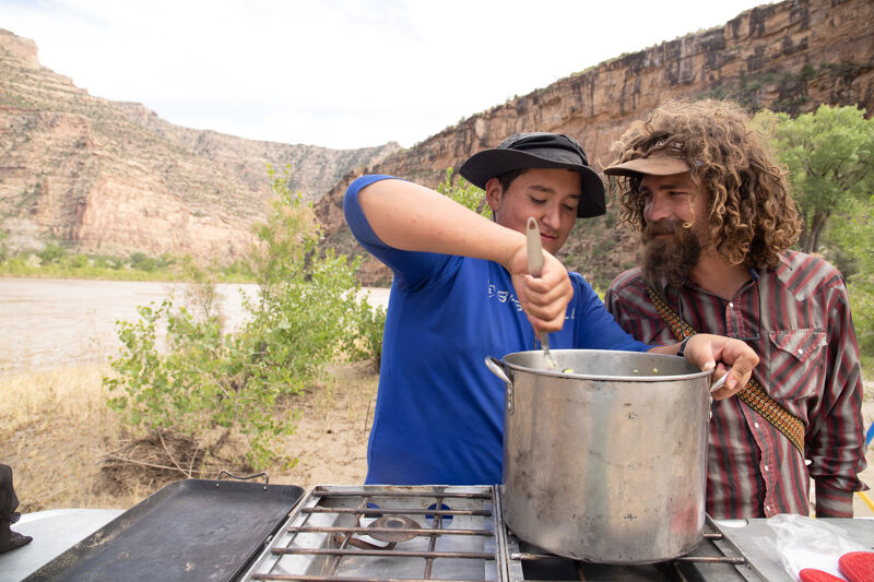 In the image, two people are cooking outdoors. One person, wearing a blue shirt and a hat, is stirring a large pot on a gas stove. The other person, with long curly hair and a beard, is watching. They appear to be camping, with a river and rocky cliffs in the background. A griddle sits next to the stove, and the overall scene suggests they are preparing a meal in a natural setting.